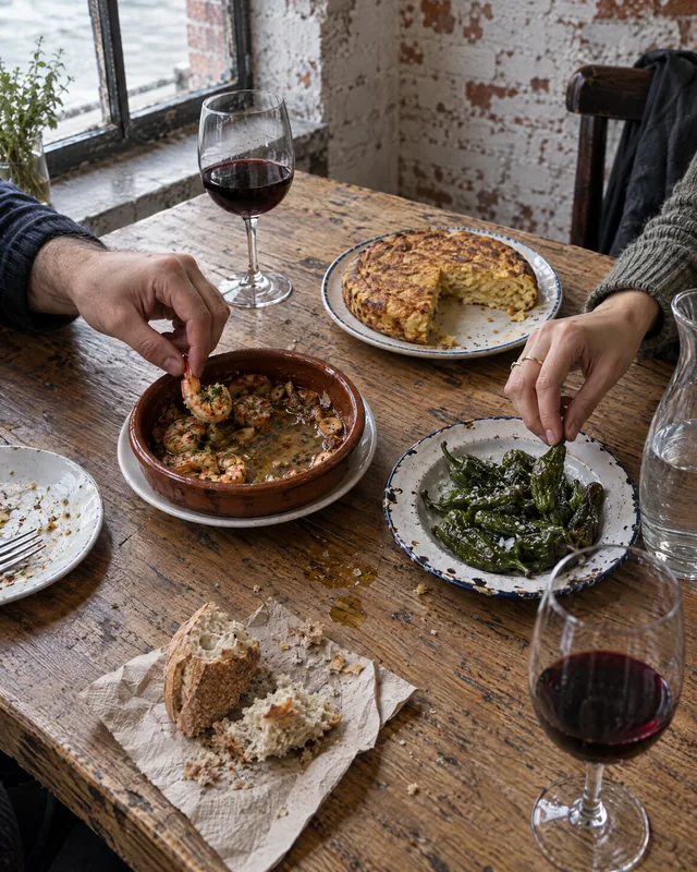 Geteilte Tapas von oben: Hände greifen nach Gambas, Padrón, Tortilla und Brot, dazu zwei Gläser Rotwein