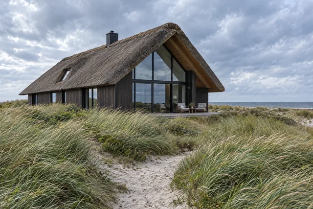 Reetdachhaus mit Glasgiebel hinter Dünengras unter bewölktem Himmel